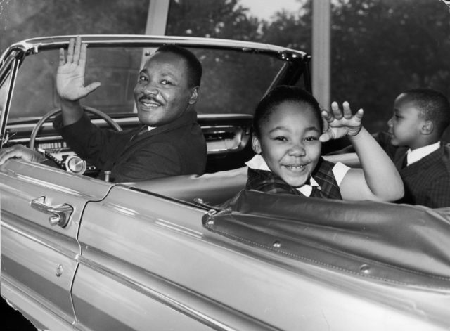 Martin Luther King Jr. looking back from the front seat of a car, his son and daughter waving