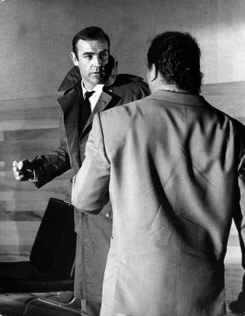 Sean Connery wears a trench coat with the collar popped, winding up a punch for a man standing in front of him.