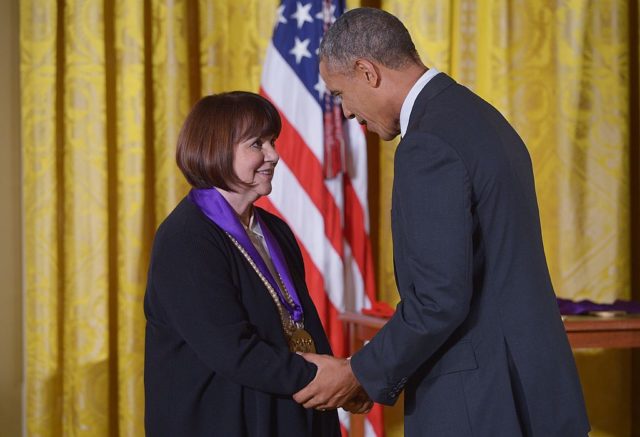 Linda Ronstadt in a black jacket with medal on her neck shakes hands with Barack Obama, also in a suit jacket.