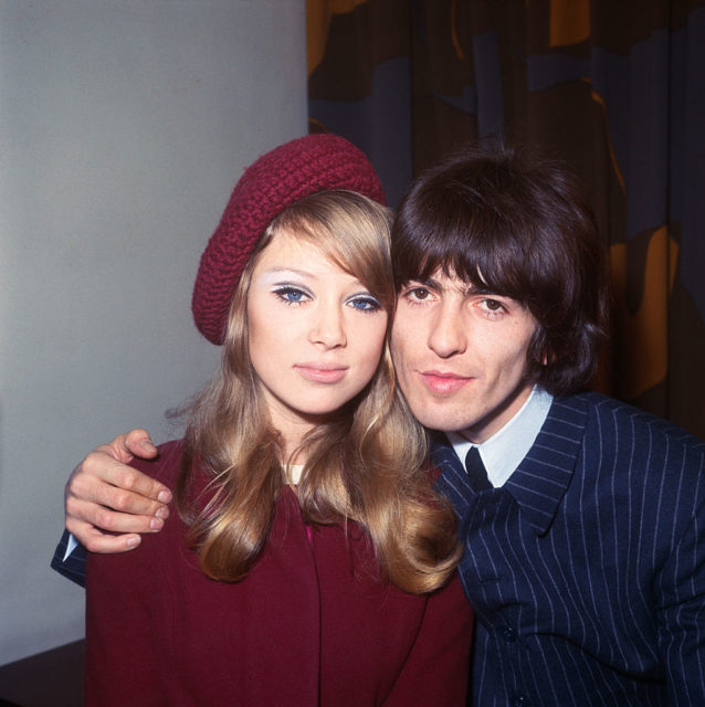 George Harrison in a blue pin stripe suit puts his arm around Pattie Boyd, wearing a red knit beret and matching jacket.