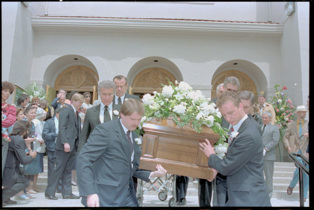 Group of male pallbearers, including Glenn Ford, carry Rita Hayworth's coffin down the steps of a church through a crowd. 