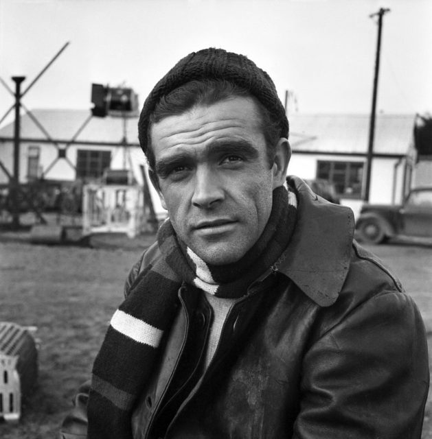 Sean Connery looking into the camera while wearing a leather jacket, striped scarf, and knitted hat.