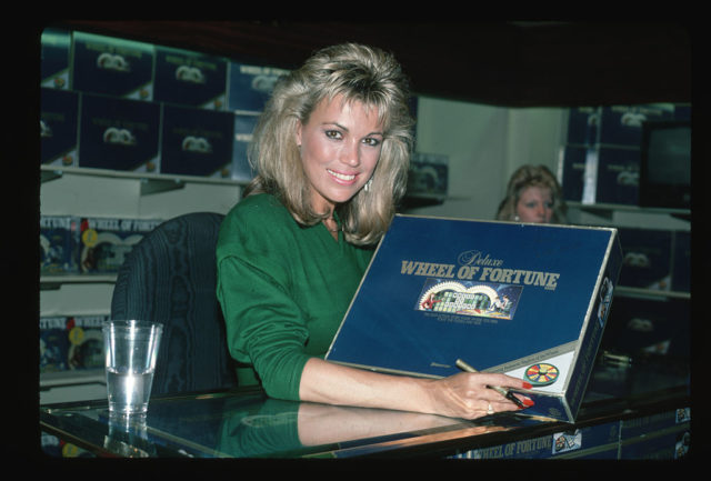 Vanna White in a green dress with a copy of a 'Wheel of Fortune' board game, posed to sign it.