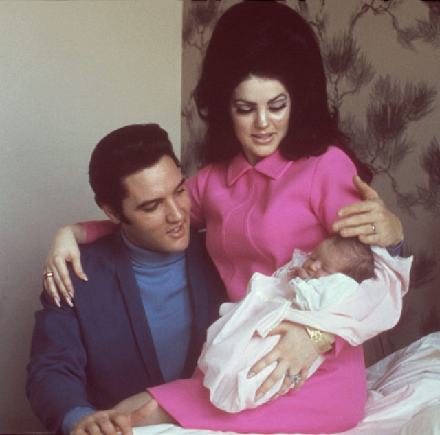 Priscilla Presley in a pink jacket dress holding a baby in a blanket with her arm around Elvis who sits beside her in a suit jacket and turtle neck.
