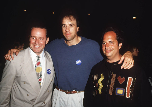 Phil Hartman, Kevin Nealon, and Jon Lovitz smiling at the camera with their arms around each other.