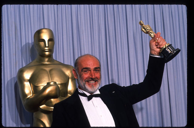 Sean Connery holds up his Oscar while wearing a black suit and bowtie in front of a life sized model of the same statue.