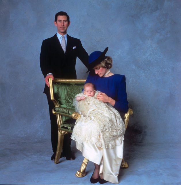 Princess Diana sitting in a chair, baby Prince Harry in her lap. Prince Charles stands behind the chair