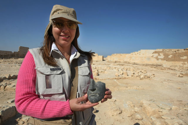 An archaeologist holds the remains of a statue's face, excavation site in the background