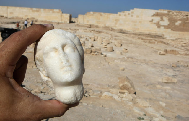 A hand holds the head of a small statue in front of an excavation site