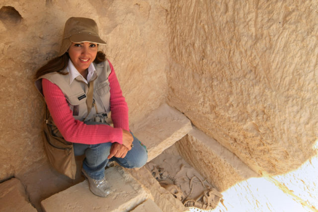 Archaeologist sitting at a hole int he ground, walls around her