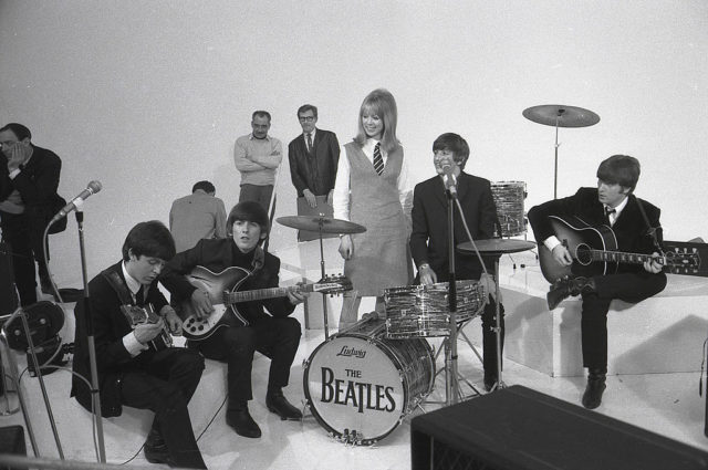 Pattie Boyd in a dress over a collared shirt and tie stands with The Beatles and their drum set.