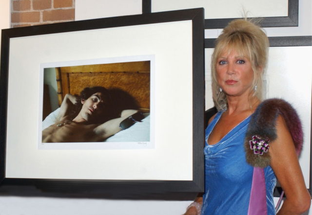 Pattie Boyd in a blue dress standing beside a framed photo of George Harrison shirtless in bed.