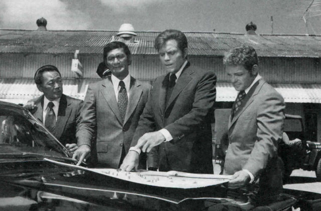 Jack Lord, Al Harrington, Kam Fong, and James MacArthur wearing suits surrounding a map sitting on a car.