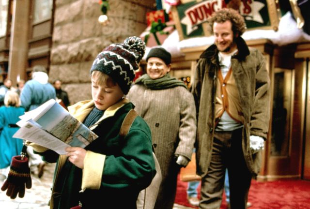 Macaulay Culkin in a hat and jacket looks down at a map while Joe Pesci and Daniel Stern stand behind him staring down.