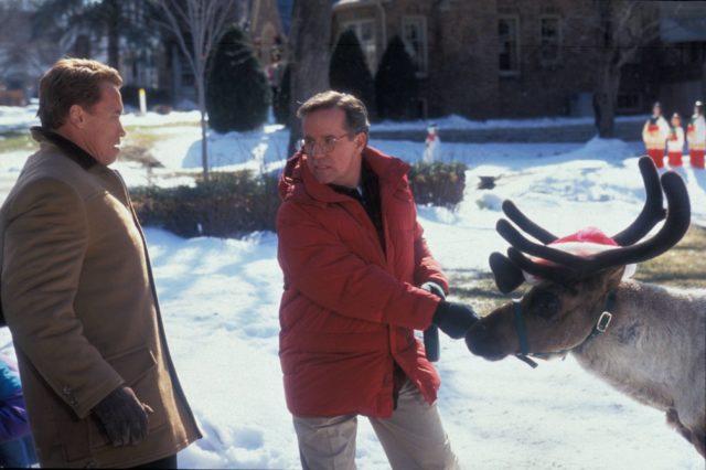Phil Hartman and Arnold Schwarzenegger wearing jackets while standing with a reindeer in the snow.