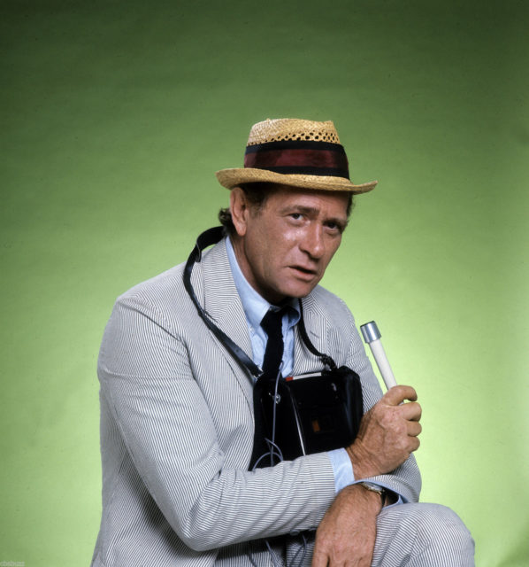 Darren McGavin as Carl Kolchak sitting in front of a green screen in a grey suit and fedora. 