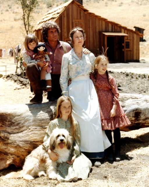 Cast of 'Little House on the Prairie' sitting on a large felled tree looking off into the distance with a cabin in the background. 