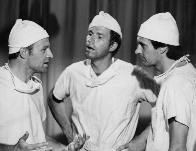 Larry Linville, Wayne Rogers, and Alan Alda as their characters wearing surgeons scrubs talking with each other.