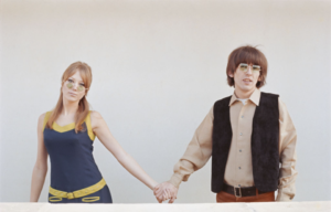 Young George Harrison with square sunglasses, a brown collared shirt and black vest, holding hands with Pattie Boyd in a blue and yellow tank top and circular sunglasses.