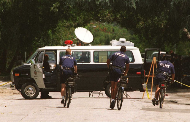 Three police officers on bicycles ride toward a police van after Phil Hartman's death