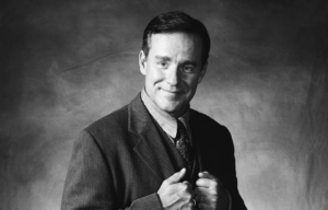 Phil Hartman wearing a suit and tie, holding the lapels of his jacket while he smiles at the camera.