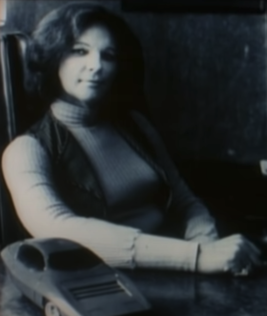 Black and white photo of Elizabeth Carmichael sitting at a desk with her arm up, beside a toy car.