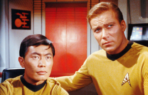 William Shatner with his arm around a seated George Takei both in yellow Star Trek uniforms.