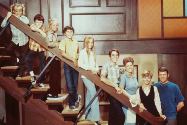 Full cast of 'The Brady Bunch' lined up on the banister down the stairs in their house, smiling at the camera. 