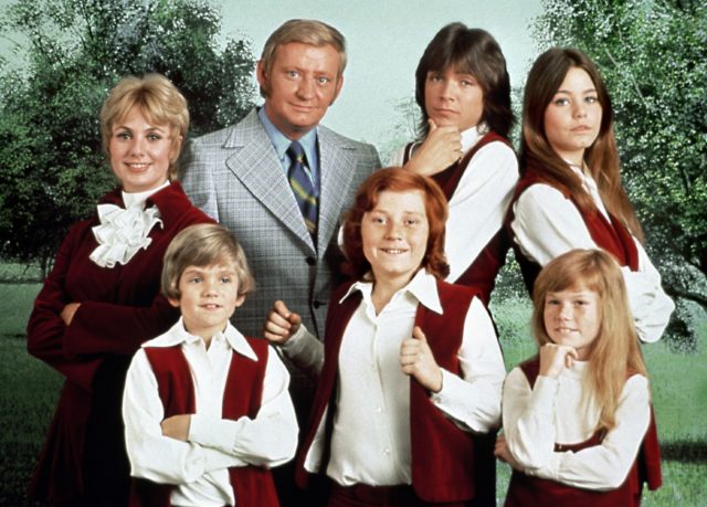 Cast of 'The Partridge Family' with the children wearing red vests and white collared shirts, while Dave Madden as Reuben Kincaid wears a grey suit, and Shirley Jones as Shirley Renfrew Partridge wears a red suit dress.