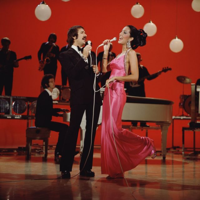 Cher wearing a long pink gown, and Sonny in a black suit, sing into a microphone together with a band in the background.