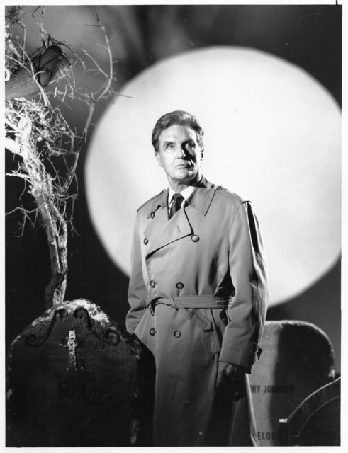 Robert Stack standing in front of a full moon beside two fake tombstones while wearing a trench coat and looking off in the distance.