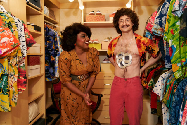 Movie still of Daniel Radcliffe as Weird Al with an open Hawaiian shirt, red pants and disks hanging from his neck, and Quinta Brunson as Oprah Winfrey in a brown dress.