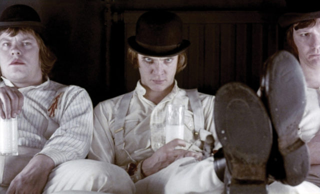 Warren Clarke, Malcolm McDowell, and James Marcus sit together in white shirts, pants, and bowler hats. 