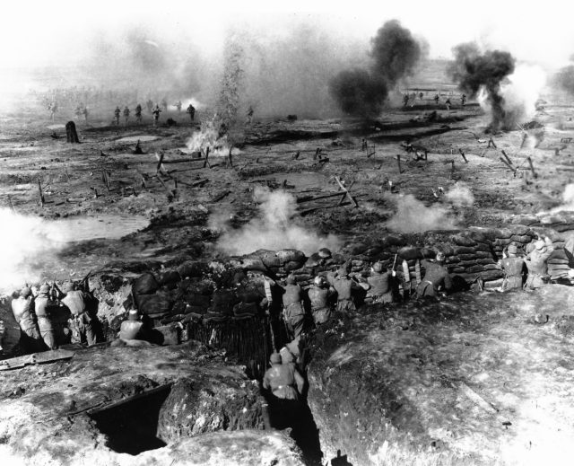 Aerial view of actors dressed as WWI soldiers firing in trenches.