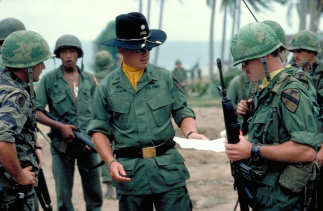 Robert Duvall in military uniform talks with a group of soldiers that surround him.
