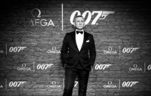 Black and white photo of Daniel Craig in a black suit with bow tie in front of a wall with "007" logos on it.