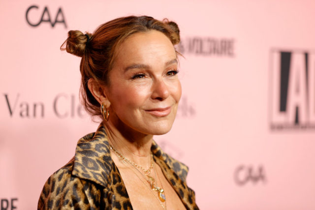 Jennifer Grey in a cheetah print shirt with her hair in two small buns smiling to the side of the camera.