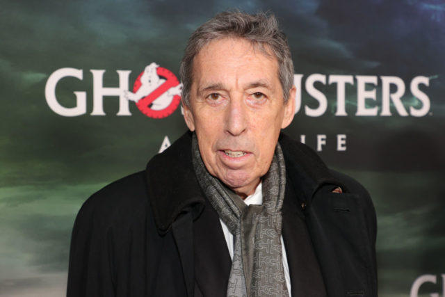 Ivan Reitman in front of a Ghostbusters background wearing a scarf and a black jacket.