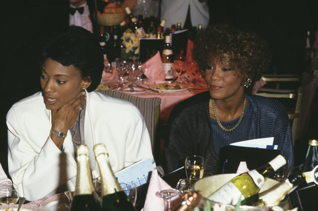 Robyn Crawford in a white jacket and Whitney Houston in an oversized top, sit side by side at a dinner table. 