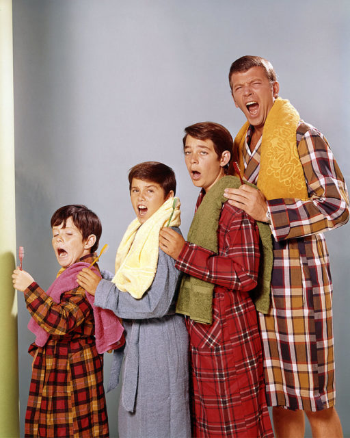 Robert Reed, Mike Lookinland, Christopher Knight, and Barry Williams stand in a row with their hands on their shoulders in house coats with towels on their shoulders. 