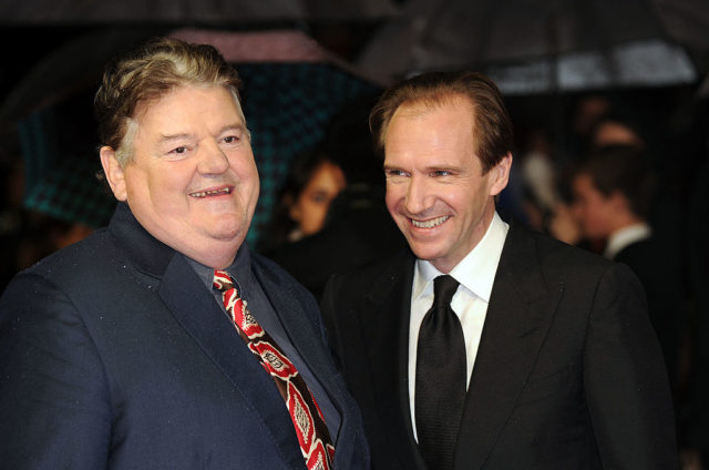 Robbie Coltrane and Ralph Fiennes in black suit jackets smiling to the press.