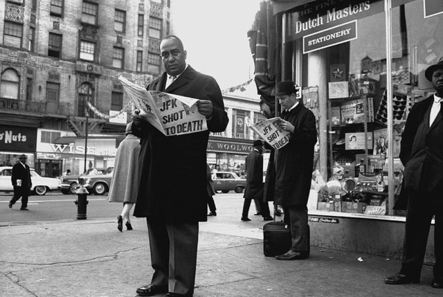 Men in black jackets stand on a street corner reading newspapers with the headline "JFK Shot to Death."