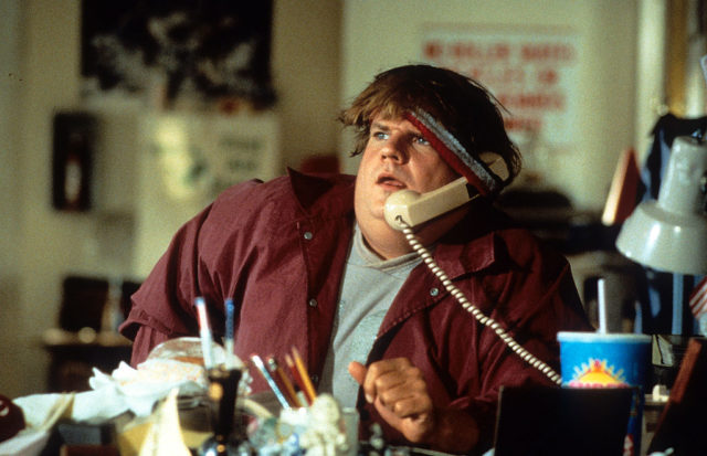 Chris Farley in a red shirt with a phone tucked under a headband holding it to his ear.