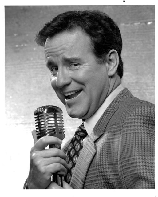 Phil Hartman in a tweed suit holding a microphone.