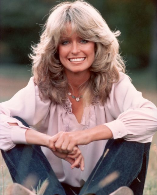 Farrah Fawcett smiling at the camera while wearing a pink shirt and jeans.
