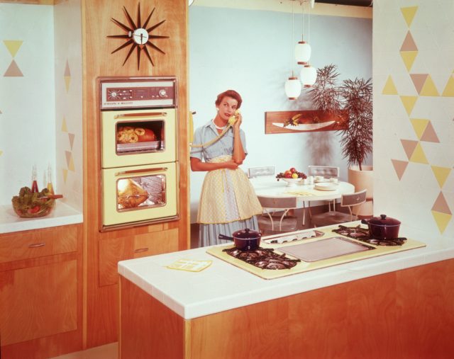 A woman speaks on the phone in her 1950s kitchen while food cooks in an oven.