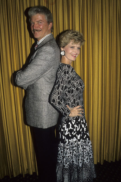 Robert Reed in a grey suit stands back to back with Florence Henderson in a black and white dress.