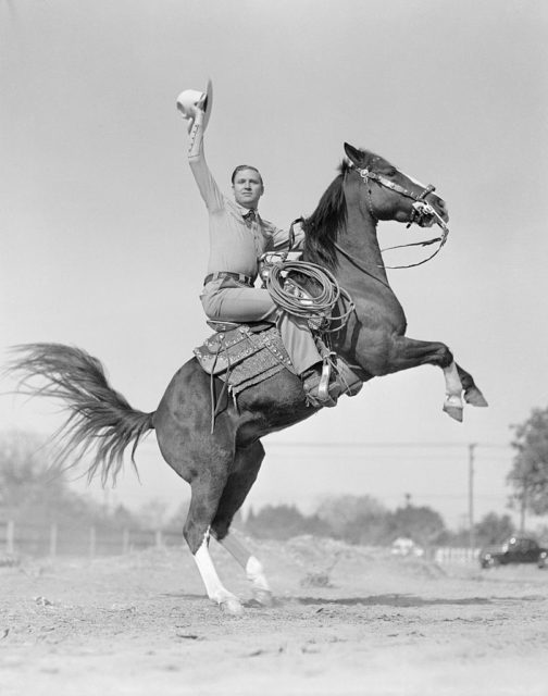 Young Gene Autry sitting on a rearing horse holding his cowboy hat in the air.