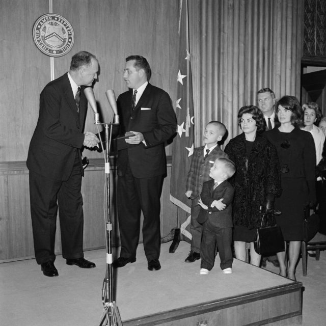 A man getting an award on stage, Jackie Kennedy and others sat to the side of the stage