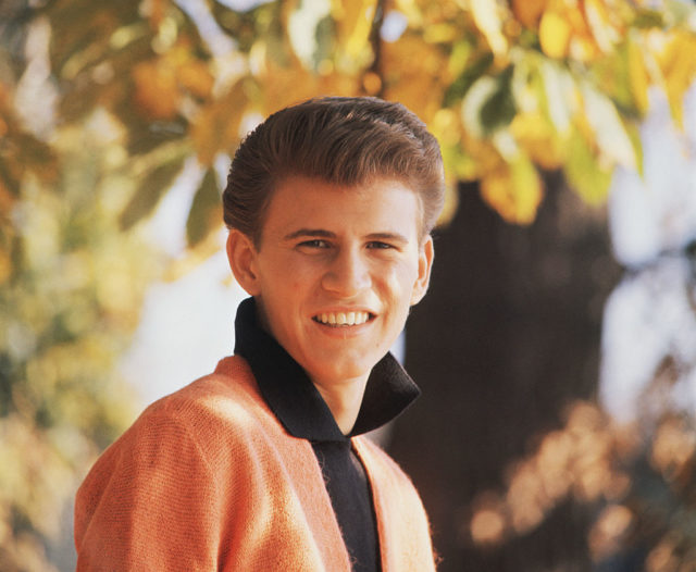 Bobby Rydell wearing an orange jacket in front of a tree. 
